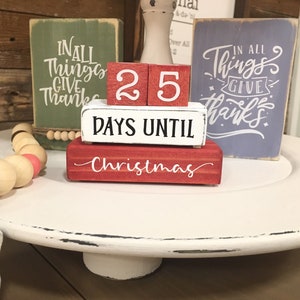 May include: A Christmas countdown block set with red, white, and green blocks. The top blocks display "25" in white on red. Below, blocks read "DAYS UNTIL" and "Christmas" in white script. Decorative wooden signs are in the background.