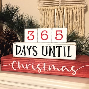 Può includere: Calendario di conto alla rovescia in legno bianco con numeri rossi che indicano "365" e il testo "DAYS UNTIL CHRISTMAS" in nero. Il calendario è su una base in legno rosso.