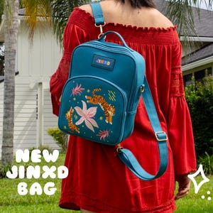 May include: A teal backpack with a floral and tiger design, worn over the shoulder. The bag has the text "JINXD" on the front and the words "NEW JINXD BAG" in a yellow font. The person is wearing a red dress.