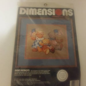 May include: A cross-stitch pattern featuring three teddy bears wearing red, white, and blue clothing and holding American flags. The pattern is titled "Bear Patriots" and is from Dimensions, a counted cross-stitch company.