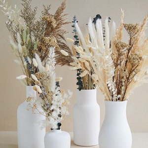 May include: Four white ceramic vases of different sizes and shapes, filled with dried flowers and grasses in shades of beige, white, and blue.
