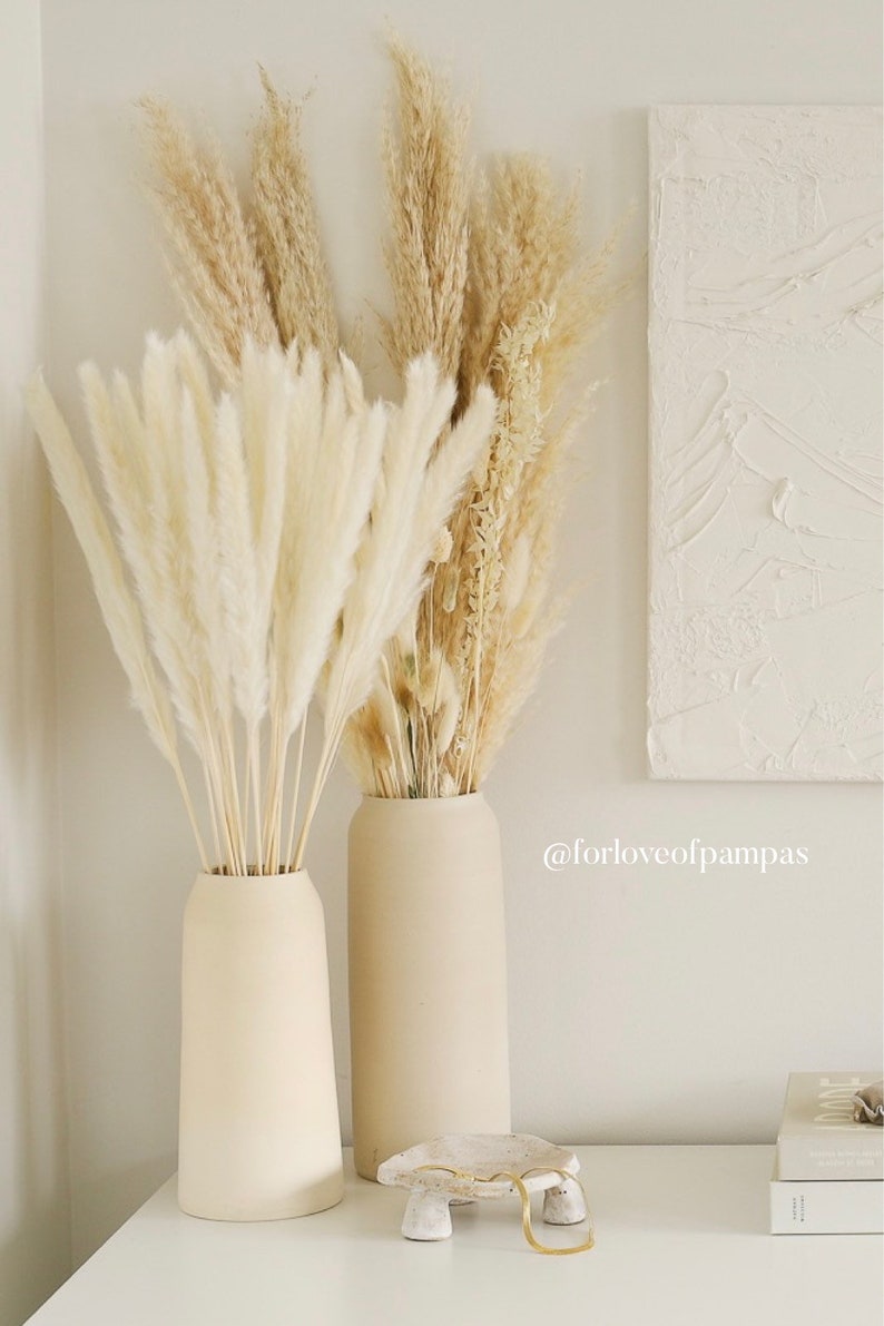 Op de afbeelding: Twee beige keramische vazen met gedroogd pampasgras in cr&egrave;me- en beigekleuren. De vazen staan op een wit oppervlak met een klein wit keramisch decoratief object met gouden accenten.