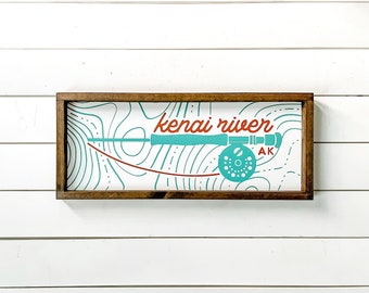 May include: A rectangular wooden sign with a brown frame. The sign features a teal fishing rod and reel design with the words "kenai river" in red and teal. The letters "AK" are also visible.