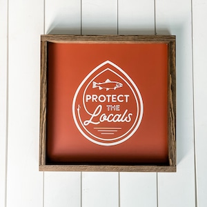 May include: A framed sign with a burnt orange background and a white graphic of a fish inside a shield shape. The text "PROTECT THE LOCALS" is displayed in white. The frame is made of dark brown wood.