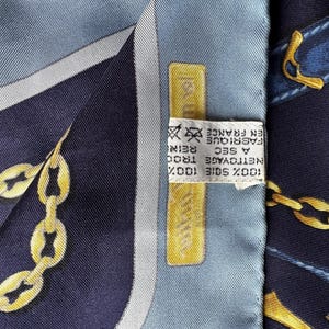 May include: A blue and white silk scarf with a gold chain design. The scarf has a label that reads '100% SOIE 100%'  'NETTOYAGE A SEC' 'FABRIQUE EN FRANCE'  'TROO REINS'.