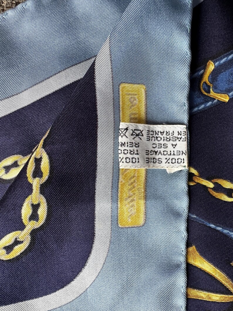 May include: A blue and white silk scarf with a gold chain design. The scarf has a label that reads '100% SOIE 100%'  'NETTOYAGE A SEC' 'FABRIQUE EN FRANCE'  'TROO REINS'.