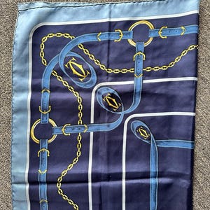 May include: A blue and gold silk scarf with a repeating pattern of chains, belts, and the word 'Cartier'.
