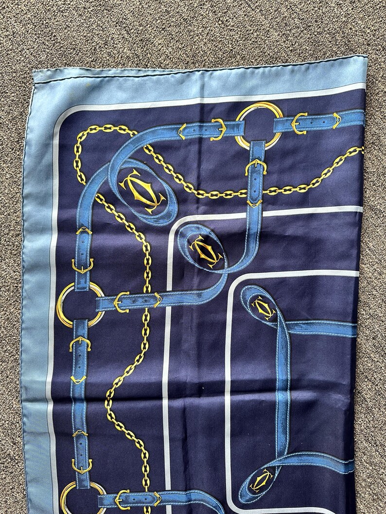 May include: A blue and gold silk scarf with a repeating pattern of chains, belts, and the word 'Cartier'.