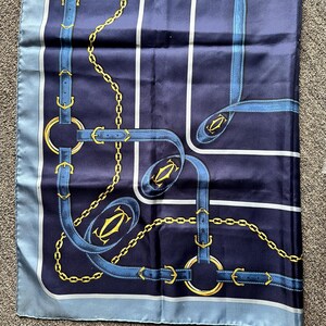 May include: A blue silk scarf with a gold chain and blue strap pattern. The scarf has a blue border and a white border. The pattern includes the Cartier logo.