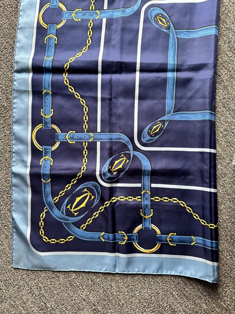 May include: A blue silk scarf with a gold chain and blue strap pattern. The scarf has a blue border and a white border. The pattern includes the Cartier logo.