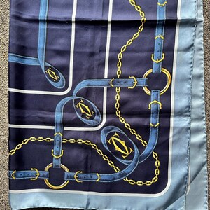 May include: A blue and white silk scarf with a gold chain and buckle pattern. The scarf has the word 'Cartier' printed on it.