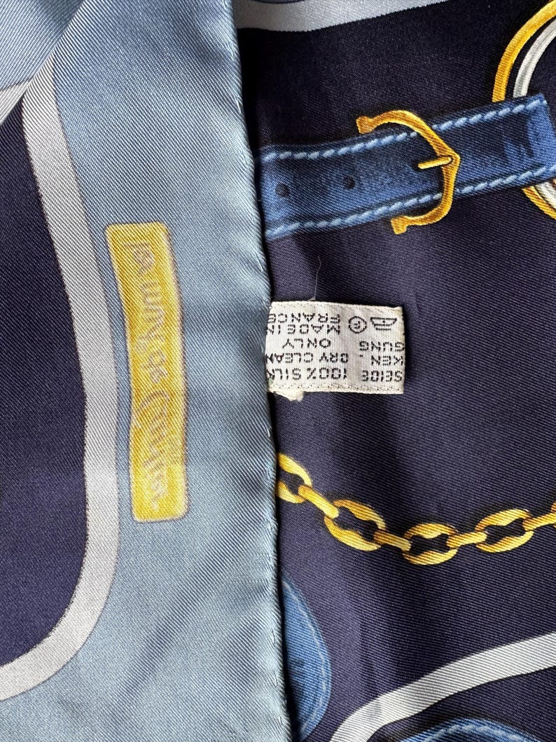 May include: A blue and white silk scarf with a gold chain and belt buckle design. The scarf has a label that reads '100% SILK - KENZO - DRY CLEAN ONLY - MADE IN FRANCE'.