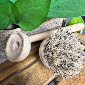 May include: Two wooden dish brushes with natural bristles. The brushes have a long wooden handle and a round, flat head. The brushes are sitting on a wooden surface.