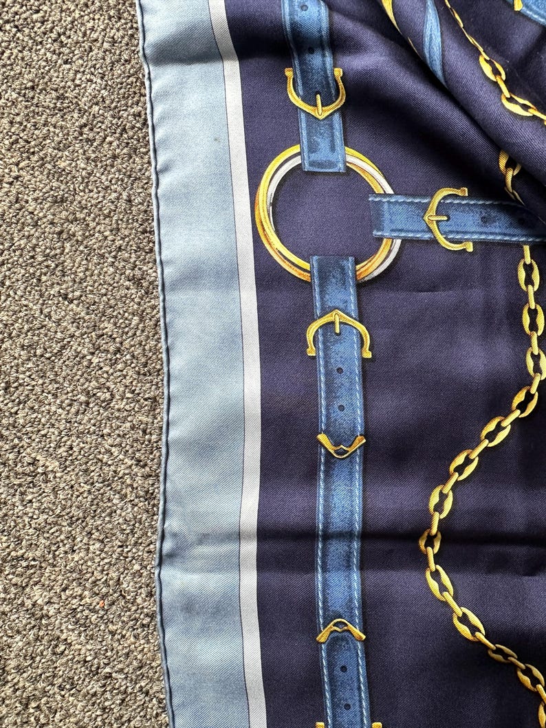 May include: A blue and white silk scarf with a pattern of gold chains and blue leather straps with gold buckles.