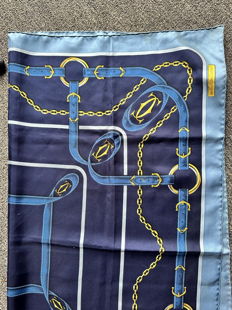 May include: A blue silk scarf with a gold chain and buckle pattern. The scarf has a light blue border and the words 'Cartier' in the top right corner.