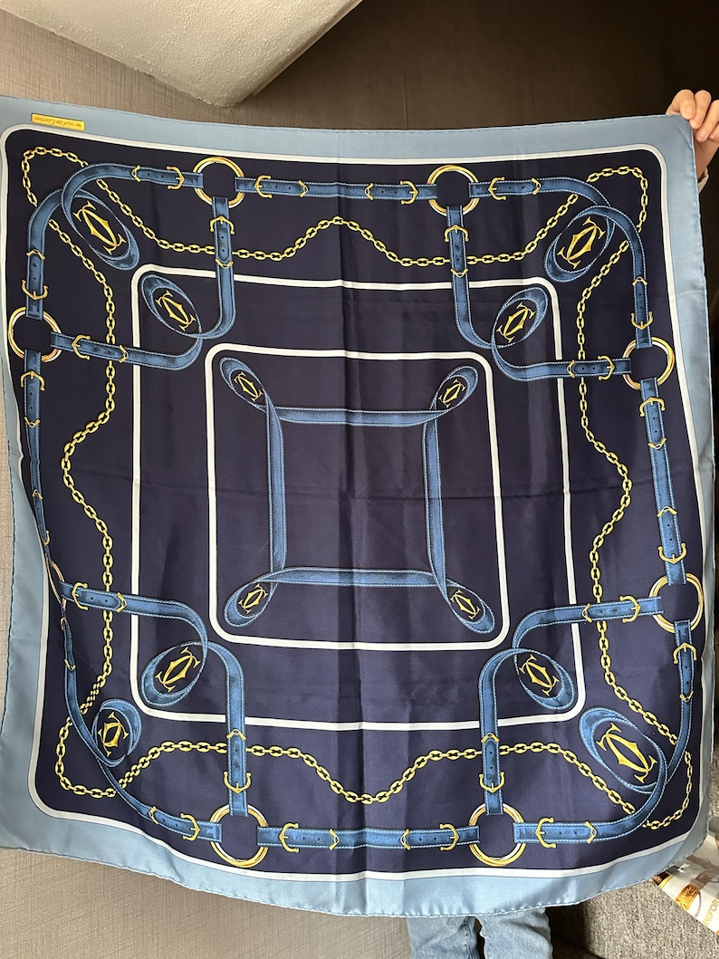 May include: A blue and gold silk scarf with a repeating pattern of chains and belts. The scarf has a light blue border and a dark blue background. The pattern is centered on the scarf and is surrounded by a white border.