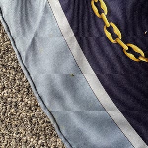 May include: A close-up of a blue and white patterned fabric with a gold chain design. The fabric has a white border and is folded over on itself.
