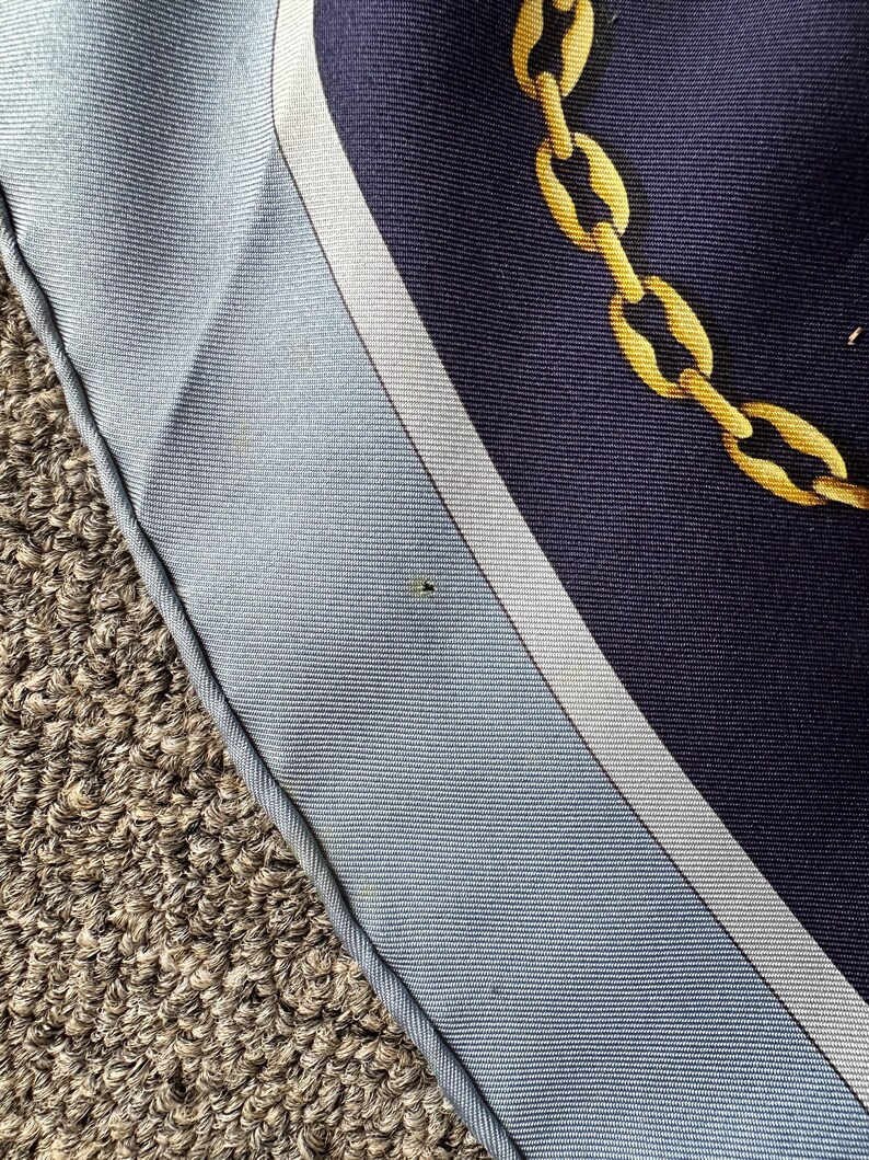 May include: A close-up of a blue and white patterned fabric with a gold chain design. The fabric has a white border and is folded over on itself.