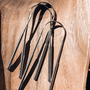 May include: Several stainless steel tongue scrapers are arranged on a wooden surface. The tongue scrapers have a curved, flattened end for scraping and a cylindrical handle. The image is lit with natural light, casting shadows.
