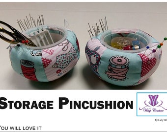 May include: Two round storage pincushions with a clear plastic insert, filled with pins, needles, and sewing tools. The fabric is patchwork with sewing-themed patterns in pink, red, and teal. The text "STORAGE PINCUSHION" is at the bottom.