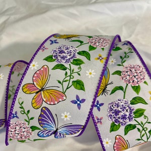Può includere: Nastro viola e bianco con un motivo floreale e farfalle. Il nastro ha un bordo viola e presenta farfalle, fiori e viti.