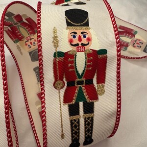 May include: Cream-colored ribbon featuring a repeating pattern of a nutcracker soldier in a red coat, black boots, and a tall black hat. The ribbon's edges are trimmed with a red stitched border, perfect for holiday crafts and decorations.