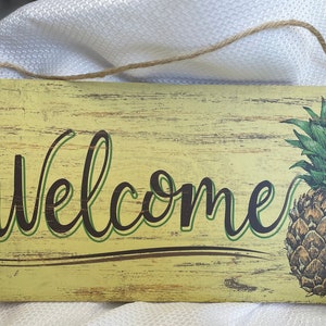 Può includere: Un cartello di legno giallo con una finitura usurata, che presenta la parola "Benvenuti" in un carattere corsivo verde e nero. Un ananas con foglie verdi è raffigurato sul lato destro del cartello.