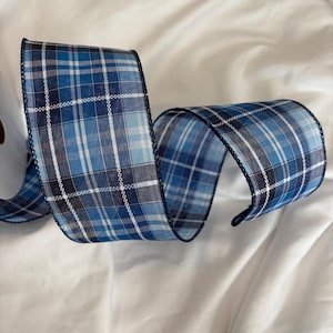 May include: A roll of blue plaid ribbon with a dark blue stitched edge. The ribbon features a pattern of light and dark blue squares with white lines. The ribbon is curled and sits on a white surface.