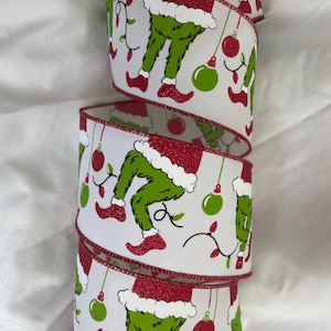 Puede incluir: Cinta blanca con ribete de purpurina roja que presenta un patrón repetido de las piernas y los pies del Grinch con botas rojas, pantalones verdes y un gorro de Papá Noel rojo con ribete blanco. La cinta también tiene adornos navideños verdes y rojos y luces blancas.