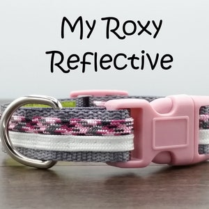 Op de afbeelding: Een grijze hondenriem met roze en witte versiering en een roze plastic gesp. De riem heeft een zilveren D-ring en de tekst "My Roxy Reflective" staat boven de riem.