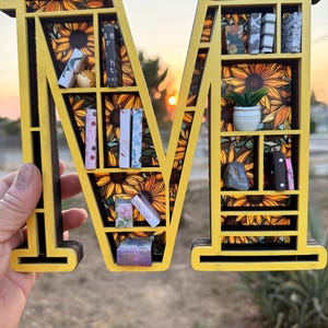 May include: A yellow wooden letter "M" with a sunflower pattern background, serving as a decorative shelf. The shelves hold miniature books, a small potted plant, and other decorative items. The letter is held up against a sunset.