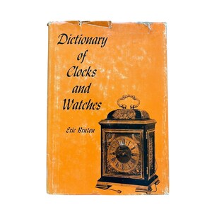 Puede incluir: Un libro naranja titulado "Dictionary of Clocks and Watches" de Eric Bruton. La portada presenta un reloj negro y dorado con números romanos.