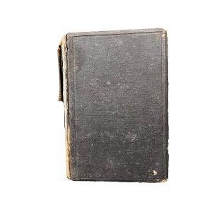 May include: A vintage, hardcover book with a worn, dark gray cover. The edges of the book show signs of age and wear, with visible discoloration and fraying. The book's cover has a textured appearance.