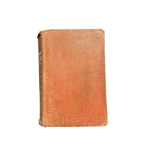 Könnte beinhalten: Ein altes Buch mit einem strukturierten, orange-braunen Einband. Der Buchrücken zeigt verblassten Text. Die Kanten des Einbands sind leicht abgenutzt, was auf das Alter hindeutet. Das Buch steht vor einem weißen Hintergrund.