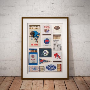 May include: A framed print featuring a collection of vintage-style matchboxes and memorabilia related to the Buffalo Bills football team. The artwork displays various logos, team colors, and illustrations of players, helmets, and a pennant on a light background.