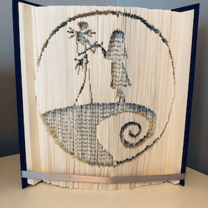Puede incluir: Una pieza de arte de libro única que presenta un diseño circular con una silueta de un personaje sosteniendo una flor. Las páginas están cortadas para crear la imagen, con un lomo azul oscuro y una cinta plateada.
