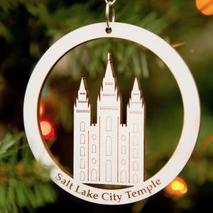 May include: A white wooden ornament shaped like a circle with a cutout of the Salt Lake City Temple. The ornament has a string for hanging and the text "Salt Lake City Temple" is written below the temple.