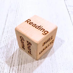 May include: A wooden cube with six sides, each with a different activity written on it: "Reading", "Screen Time", "Game", "Board Game", "Outside", and "Time".