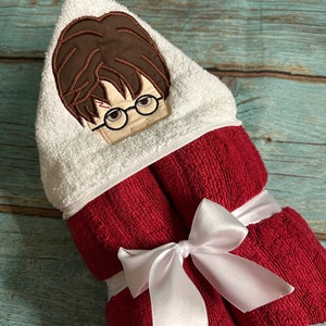 May include: A hooded bath towel featuring an embroidered design of a character with brown hair, glasses, and a lightning bolt scar. The towel is white with a red body, tied with a white satin ribbon. A fun and functional item for bath time.