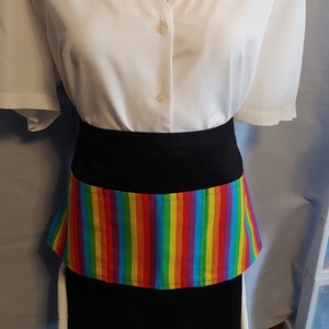 May include: A black apron with a rainbow striped pocket. The apron is worn over a white button-down shirt.