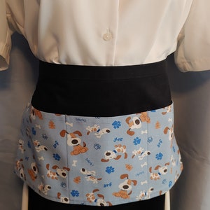 May include: A black apron with a blue and brown dog print. The print features dogs with the words "bark!" and "arf!" on a light blue background.