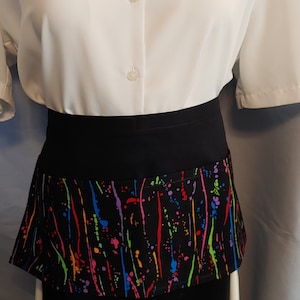 May include: Black apron with a colorful paint splatter design. The apron has a pocket on the front.