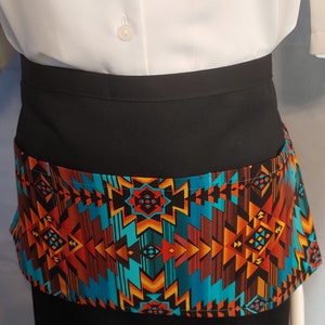 May include: A black waist apron with a colorful geometric pattern in shades of turquoise, orange, and brown. The apron is worn over a white shirt and black skirt. The apron has a pocket.