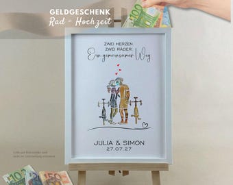 May include: A white framed print featuring a graphic of two cyclists with the German text ZWEI HERZEN. ZWEI RÄDER. Ein gemeinsamer Weg. JULIA & SIMON 27.07.27. The design includes three small red hearts above the cyclists and a heart outline below.