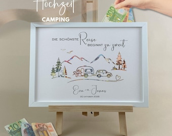 May include: A white framed sign featuring a camping illustration with a car, a caravan, mountains, and trees. The text reads "DIE SCHÖNSTE Reise BEGINNT zu zweit", "Eva ~ Jonas", and "20 OKTOBER 2026". The frame is displayed on a small wooden easel.