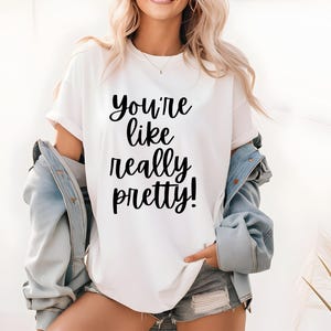 May include: White t-shirt with the phrase "You're like really pretty!" in black script. The shirt is worn with a light blue denim jacket and denim shorts.