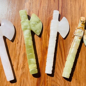 May include: Four hand-carved stone axes, each with a different color and pattern. The axes are made of white, green, and brown stone. They are all about 15 inches long.