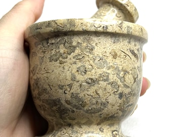 May include: A beige marble mortar and pestle with a speckled pattern. The mortar has a wide base and a rounded bowl. The pestle is cylindrical with a rounded top.