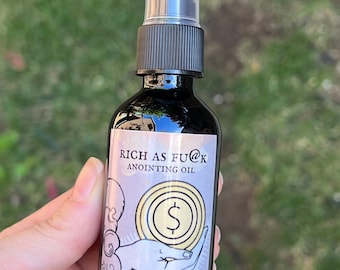 May include: A dark glass bottle with a black spray top and a label that reads "RICH AS FU@K ANOINTING OIL". The label also features a hand holding a dollar sign inside a circle.