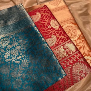 May include: Close-up of three patterned fabrics in teal, red, and orange. Each fabric features gold floral and paisley designs. The fabrics are folded, creating a textured appearance. The image showcases the rich colours and detailed patterns.